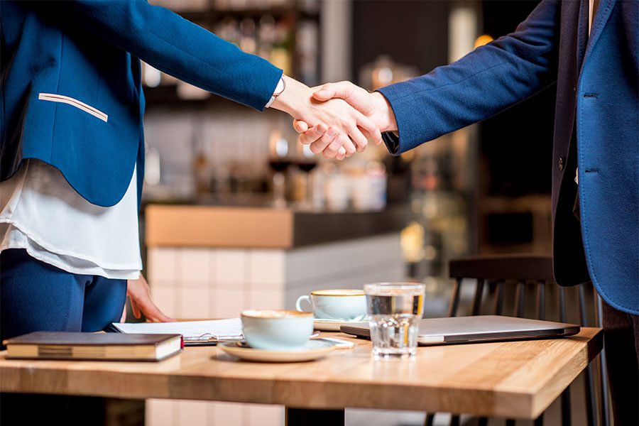 About Our Agency - Closeup View of a Business Woman and Man Shaking Hands Over a Small Table in a Restaurant with Coffee Cups