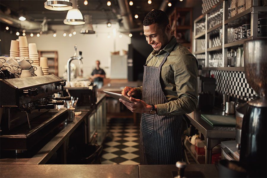 Client Center - Portrait of a Smiling Young Restaurant Owner Standing Behind the Front Counter While Using a Tablet
